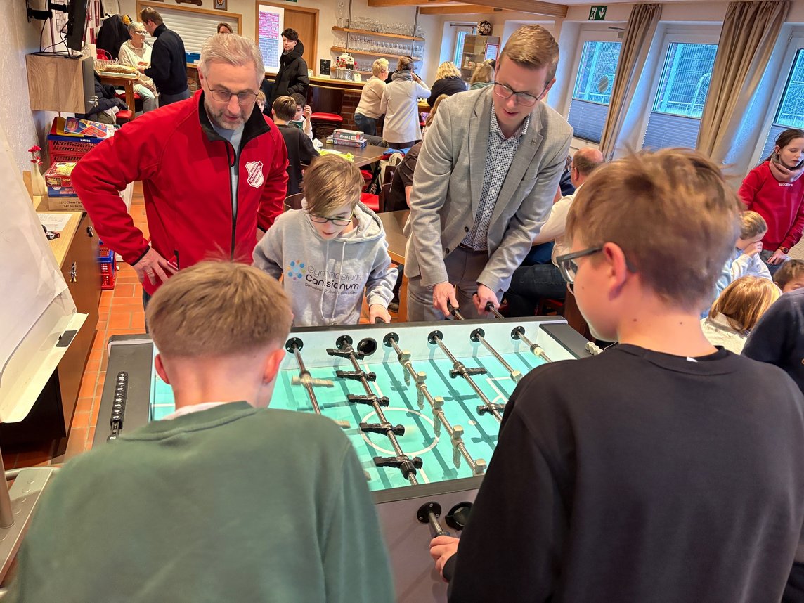 Das Bild zeigt insgesamt fünf Personen welche an einem Tischkicker spielen. 