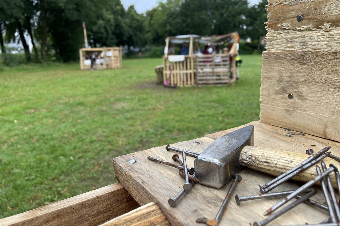 Das Bild zeigt im Vordergrund Hammer und Nägel und im Hintergrund gebaute Holzhütten.