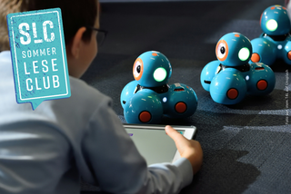 Ein Junge liegt auf dem Boden und hält ein Tablet in der Hand. Vor ihm stehen 3 "Dash" Roboter.