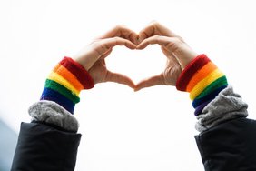 Gay Pride Heart Love Wristband Gesture Das Bild zeigt ein aus zwei Händen geformtes Herz. An den Handgelenken sind Regenbogenfarben zu sehen.