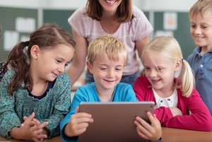 Das Bild zeigt Kinder in einer Schulklasse, die sich gemeinsam mit einer Lehrerin etwas auf einem Tablet anschauen.