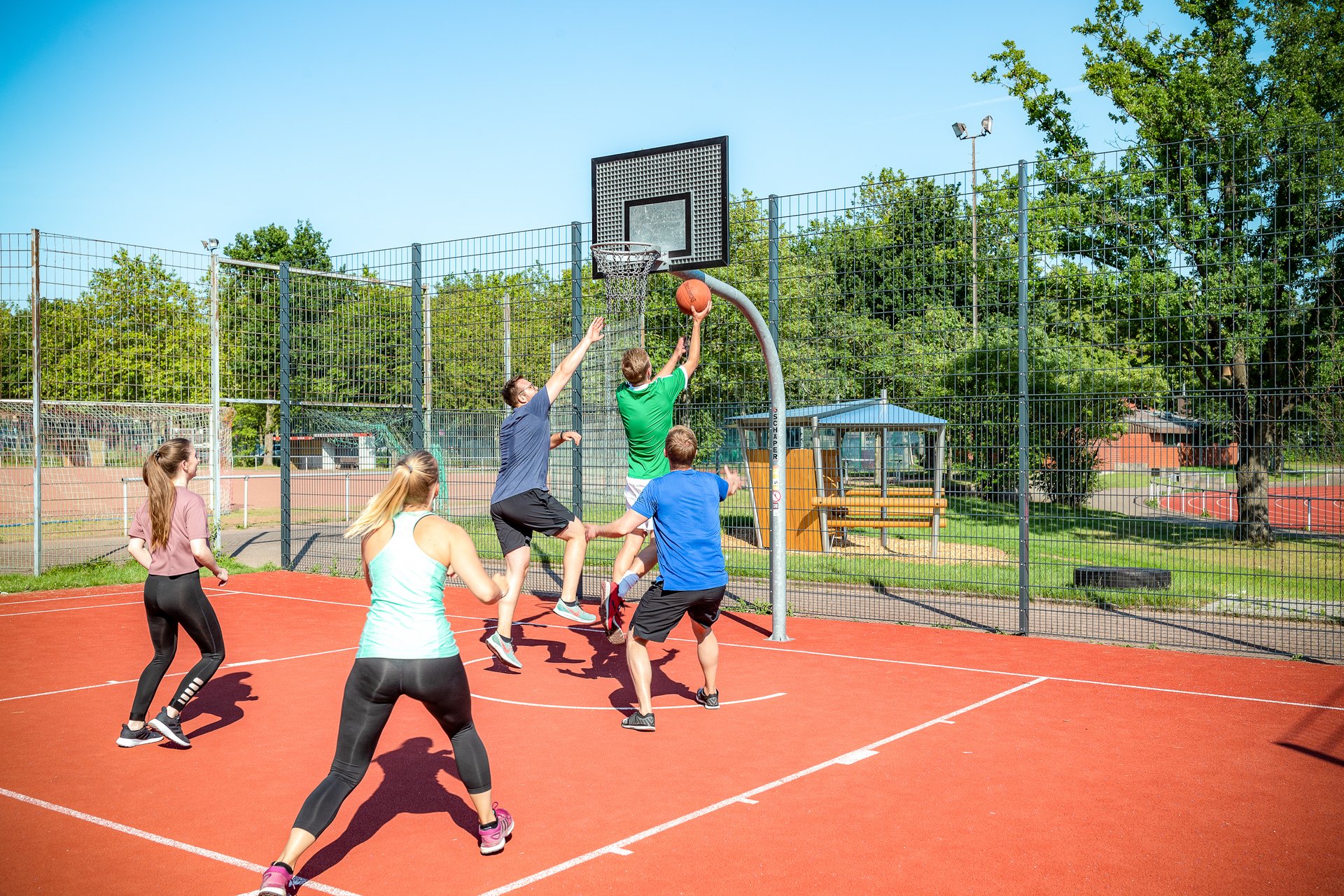 Das Bild zeigt junge Menschen, die auf eine Sportanalage im Freien Basketball spielen.